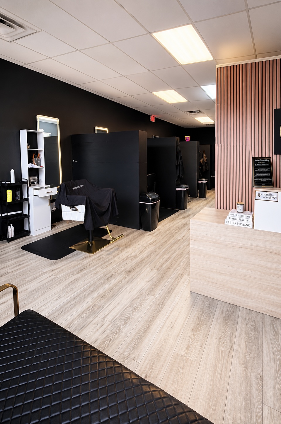 Salon with black partitions, chairs, and a reception desk, featuring wood flooring and a pink striped wall.
