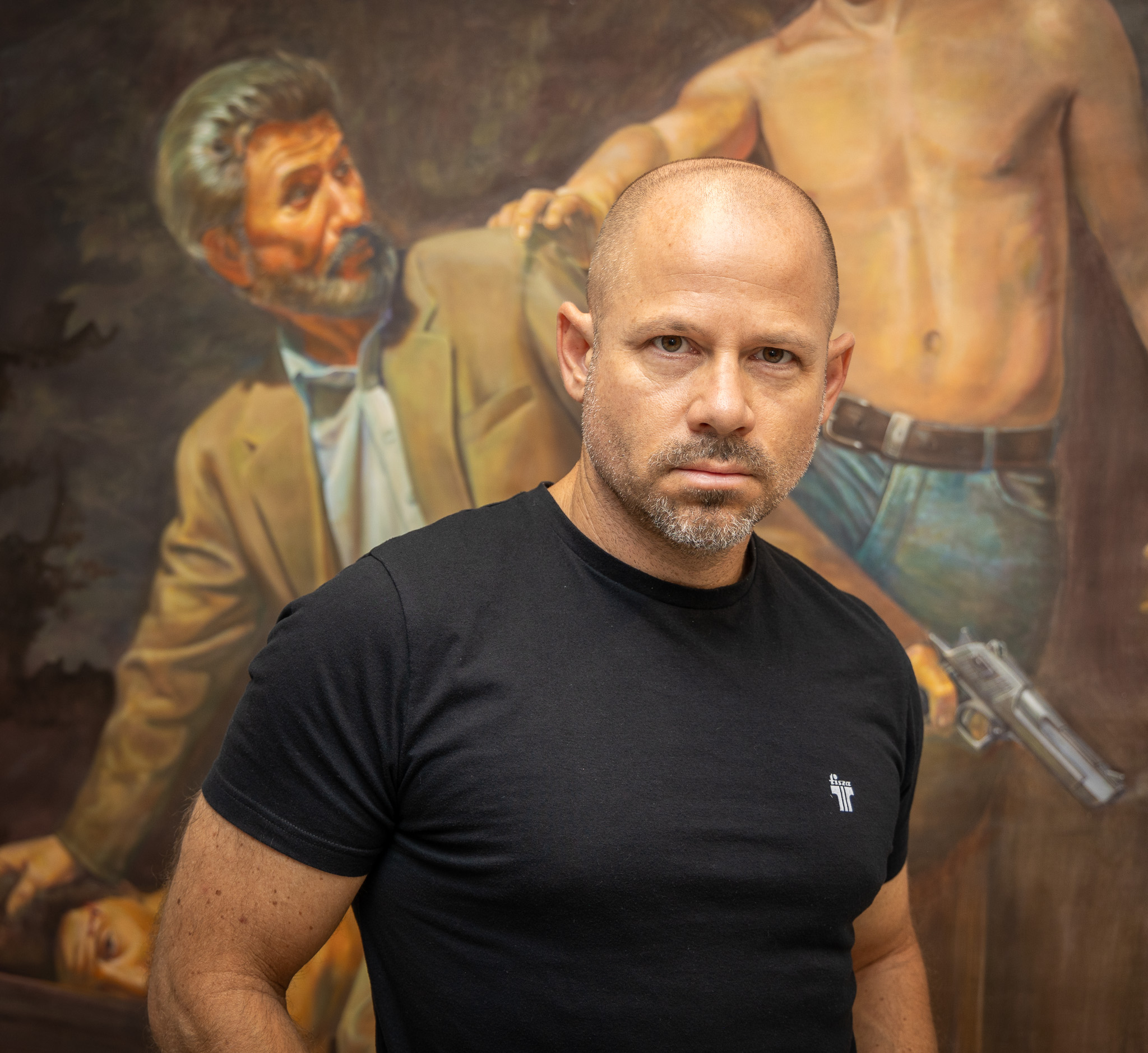 Bald man with a beard wearing a black T-shirt standing in front of a painting of a shirtless man holding a gun and a man in a suit.