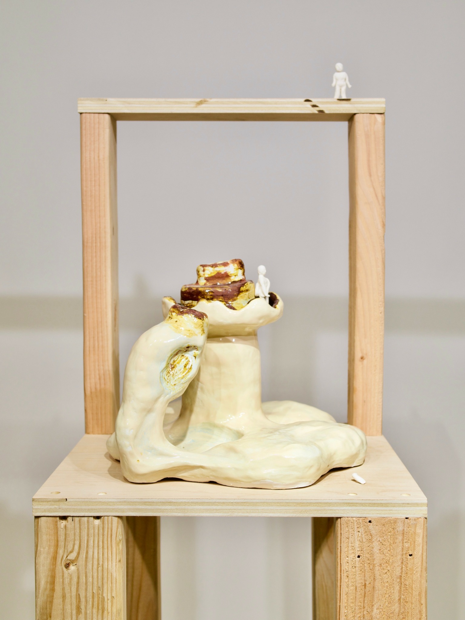 Sculpture of a person lying down with a small figure on top, framed by a wooden structure, against a plain background.