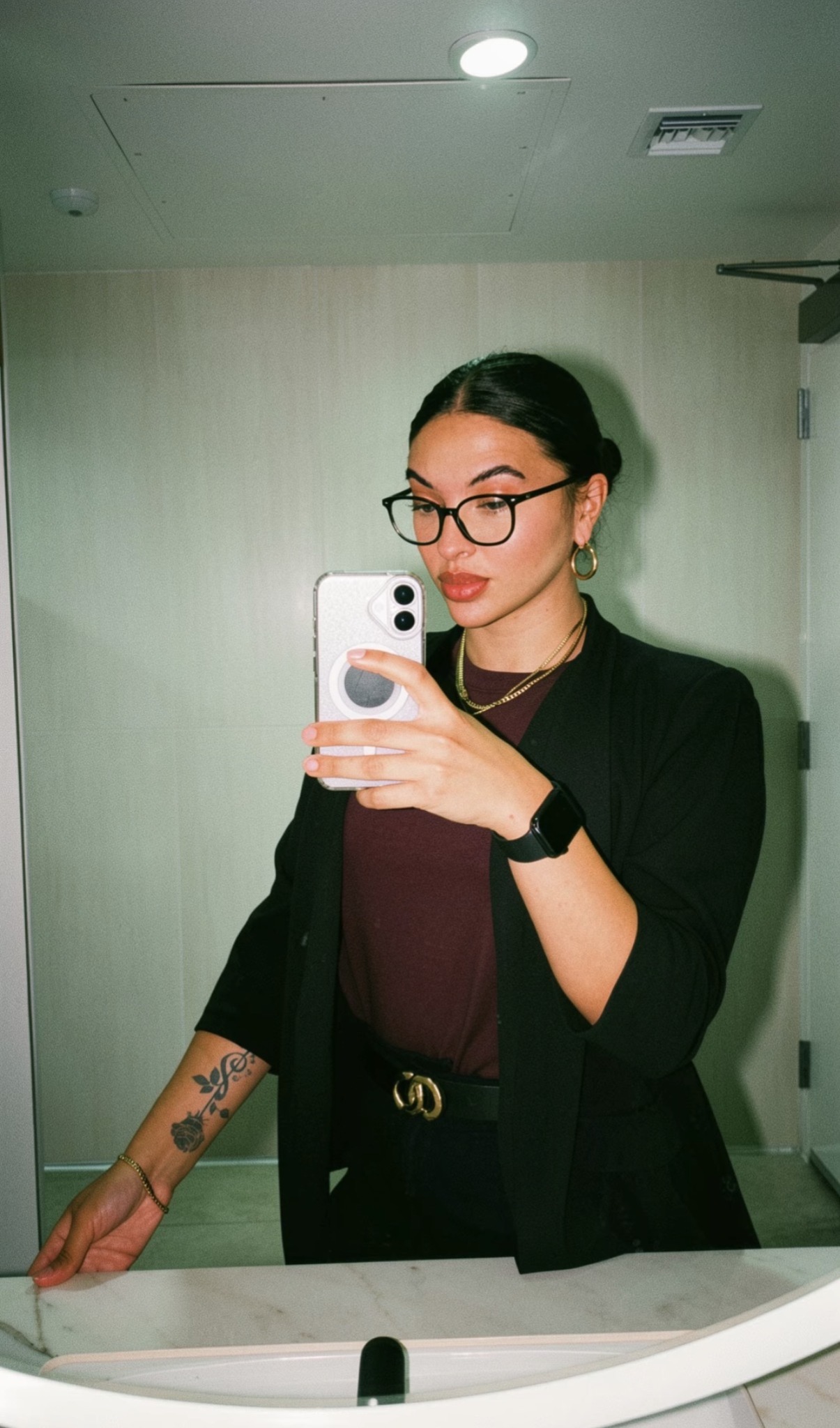 Woman with glasses and dark hair taking a mirror selfie, wearing a black blazer and maroon top, in a room with light-colored walls.
