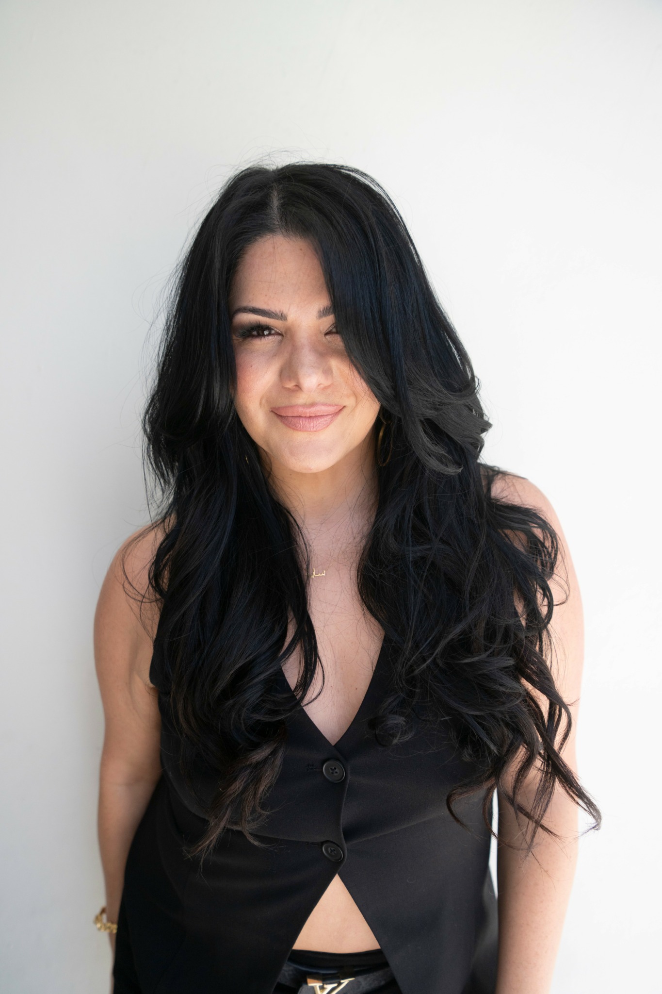Young woman with long, wavy black hair smiling, wearing a black sleeveless top, standing against a plain white background.