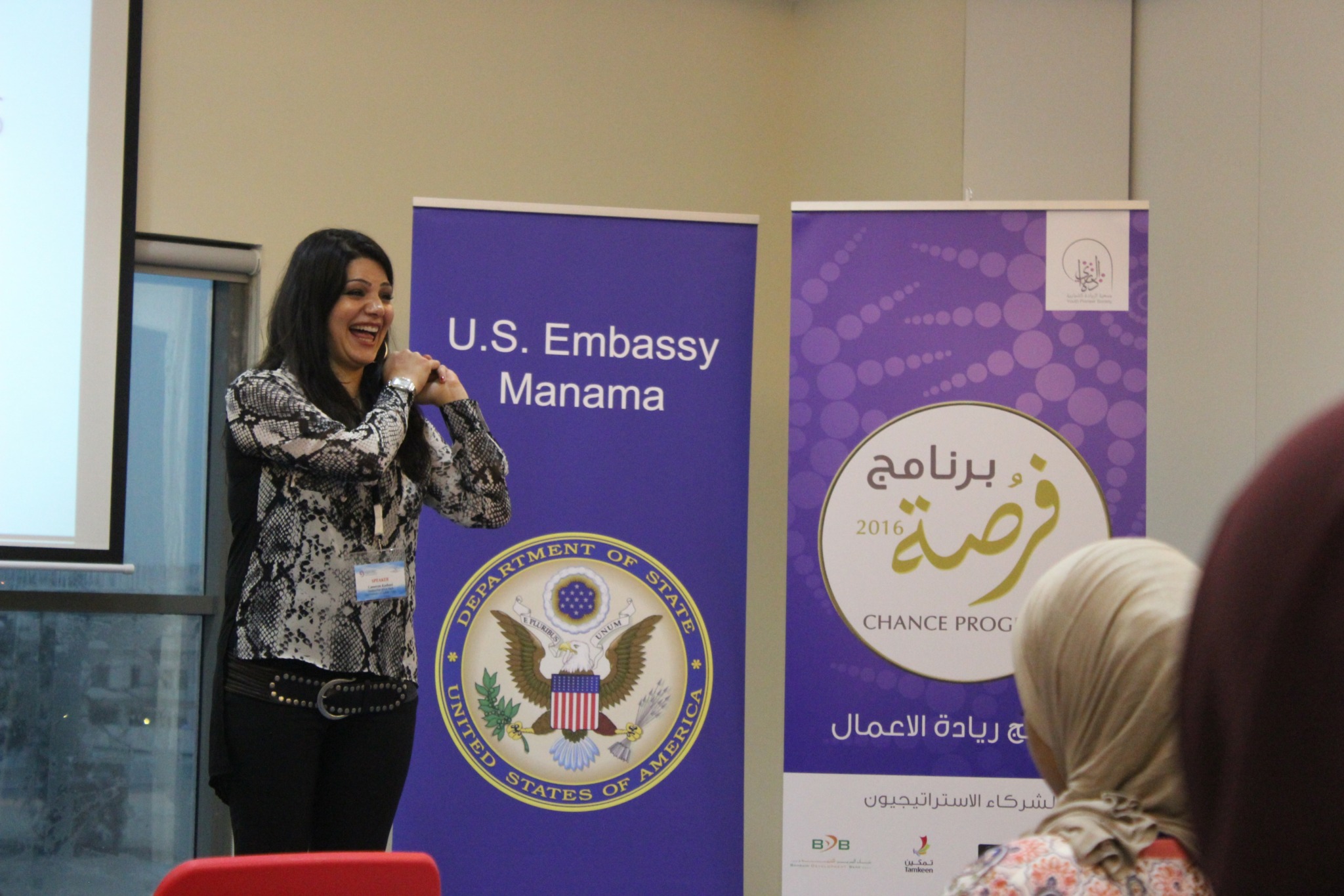 Woman speaking on stage with U.S. Embassy Manama banner and a purple event poster behind her, audience in foreground.