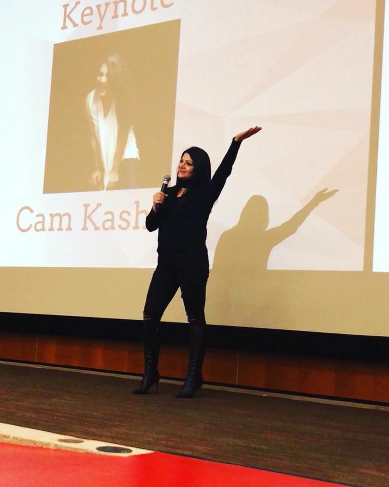 Woman speaking on stage with a microphone, gesturing with her right arm, shadow cast on screen behind her, presentation slide visible.