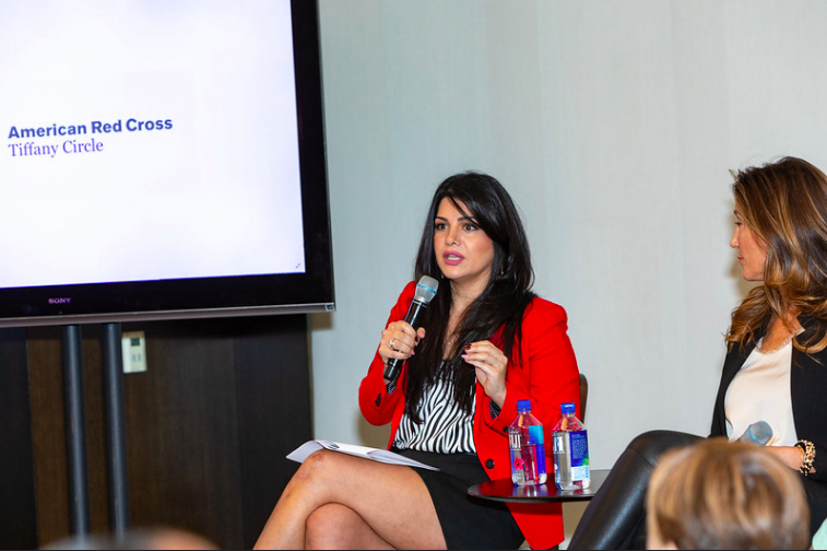 Woman in red blazer speaking into microphone during panel discussion, with screen displaying 'American Red Cross Tiffany Circle' in background.