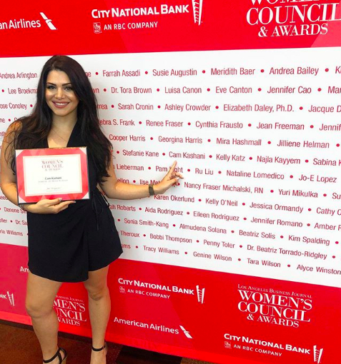 Woman in black dress holding a certificate, smiling, standing in front of a red backdrop with event logos and names.