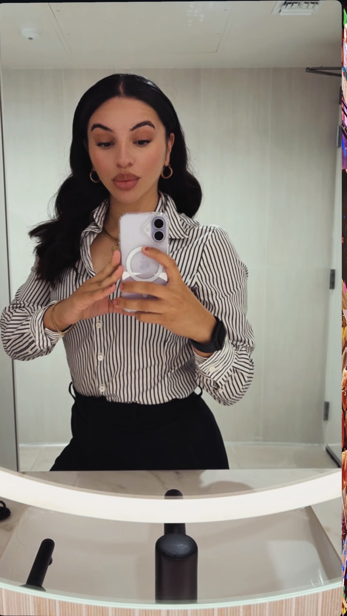 Woman taking a mirror selfie with a smartphone, wearing a striped shirt and hoop earrings, in a room with light-colored walls.