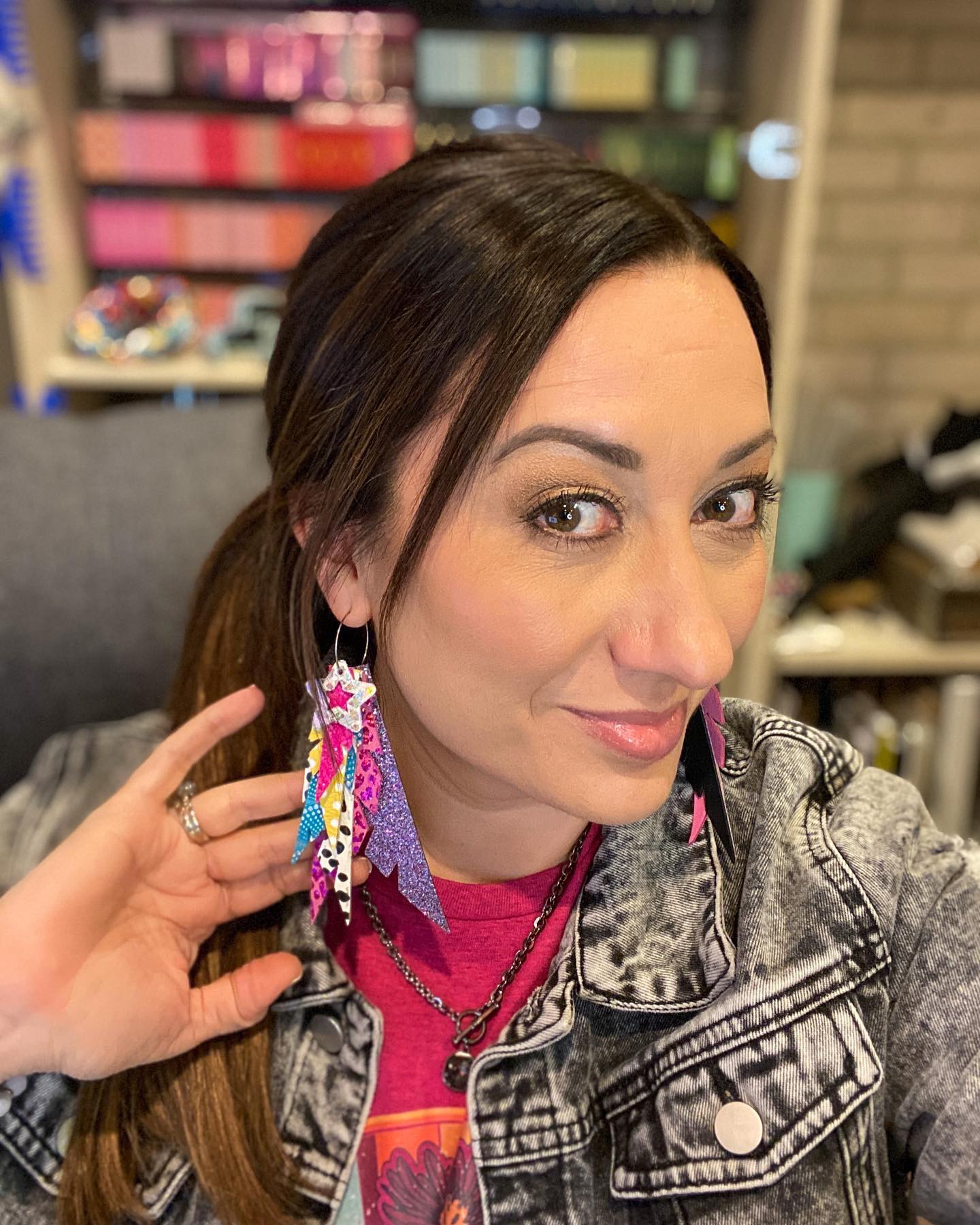 Woman with long brown hair, wearing colorful earrings and a denim jacket, sitting indoors with a blurred background.