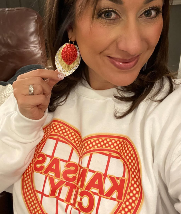 Woman smiling, holding a strawberry-shaped earring, wearing a white sweatshirt with red and orange text, indoors.