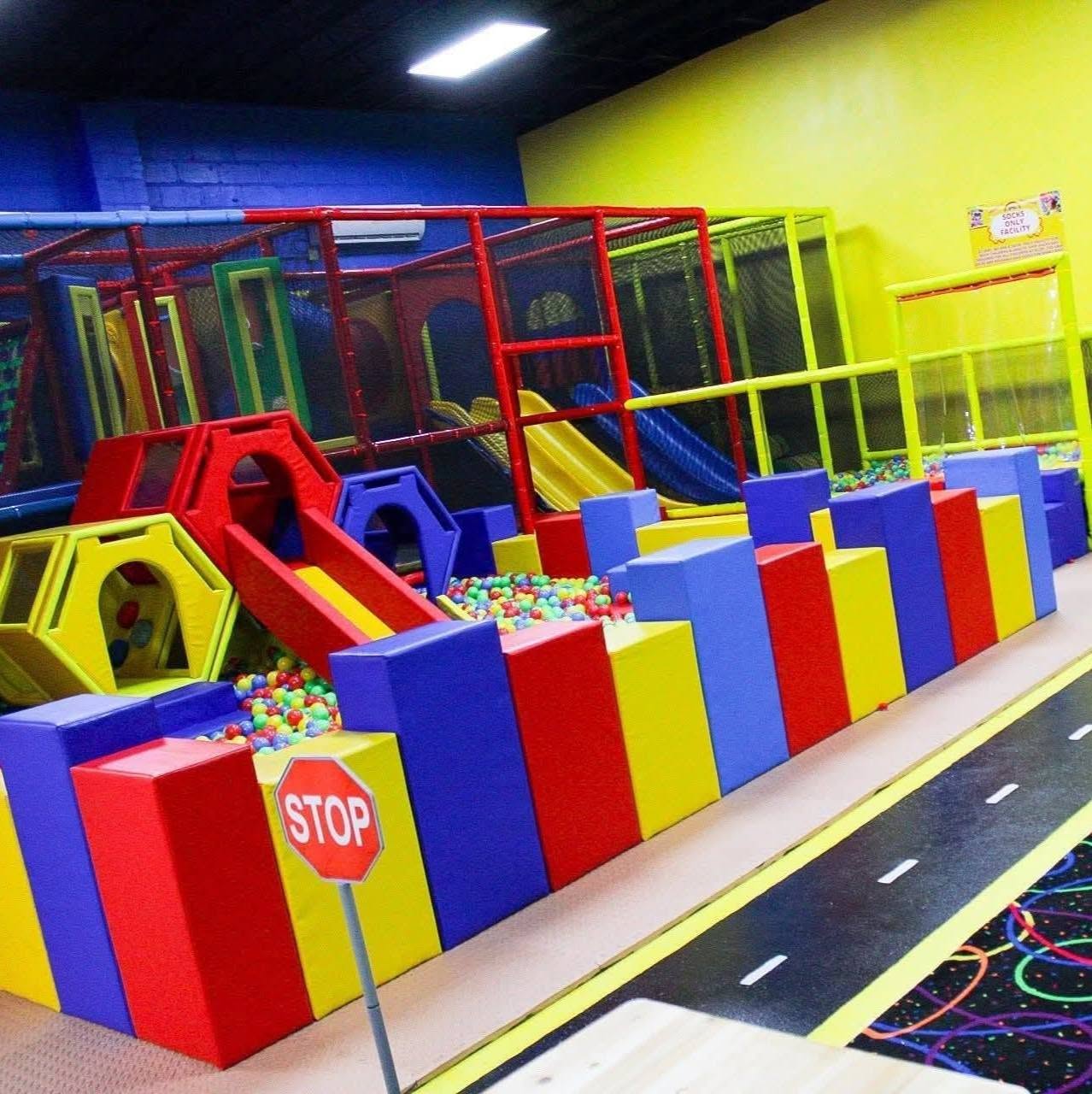 Indoor children's play area with colorful slides, ball pit, and padded barriers, against a yellow and blue wall.