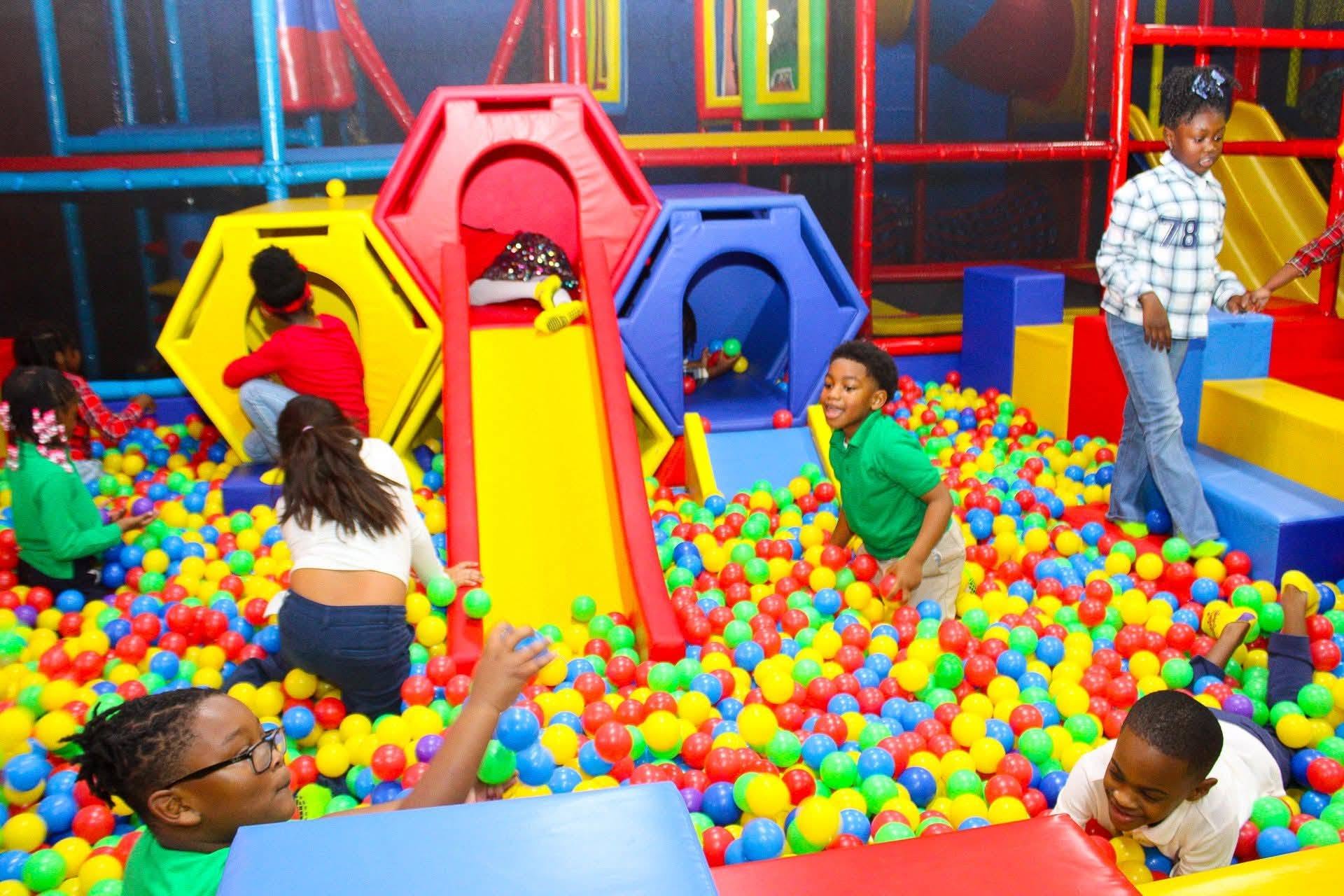 Children playing in a colorful ball pit with slides and climbing structures, adults supervising, bright indoor play area.