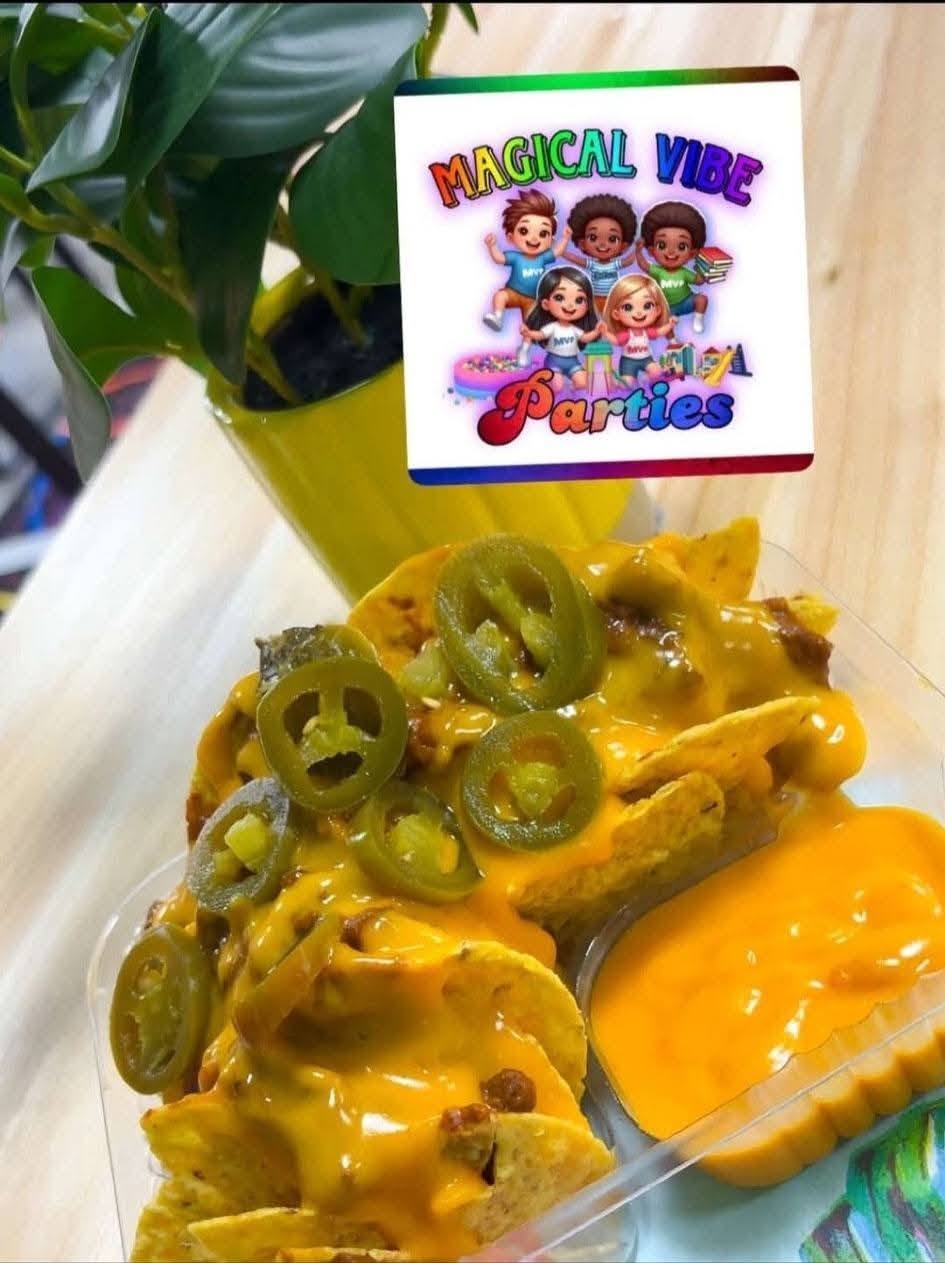 Plate of nachos with melted cheese and sliced green jalapeños, with a small bowl of cheese sauce on the side, in front of a plant with a colorful sign reading 