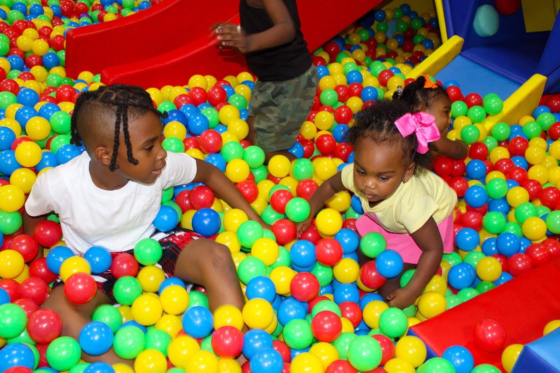 Two children playing in a colorful ball pit with red, yellow, green, and blue balls, and a third child in the background.
