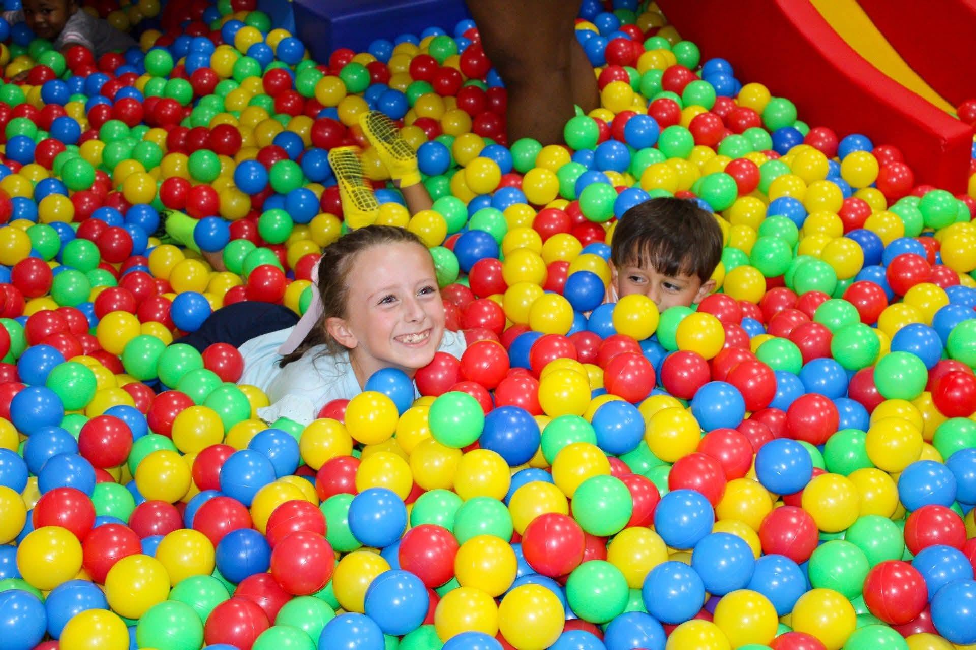 Two children are playing in a colorful ball pit filled with red, yellow, green, blue, and turquoise balls, smiling and having fun.
