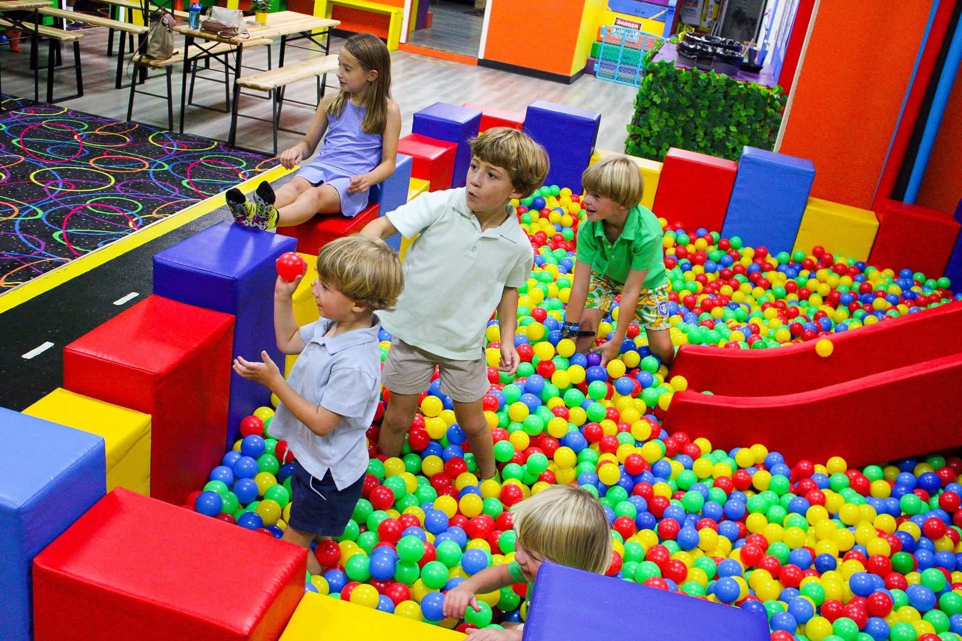 Children playing in a colorful ball pit with large foam blocks and a girl sitting nearby, in an indoor play area.