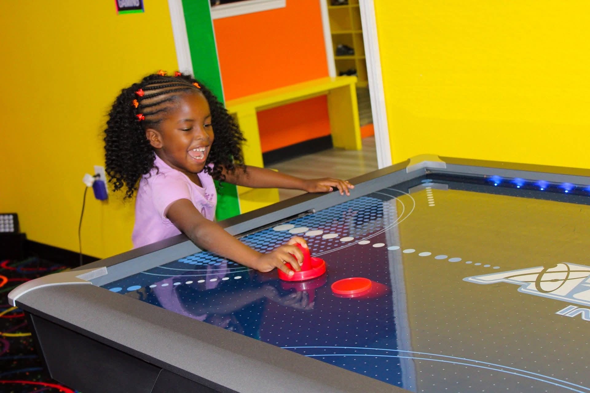 Child playing air hockey game with a red puck, smiling, in a colorful room with yellow and green walls.