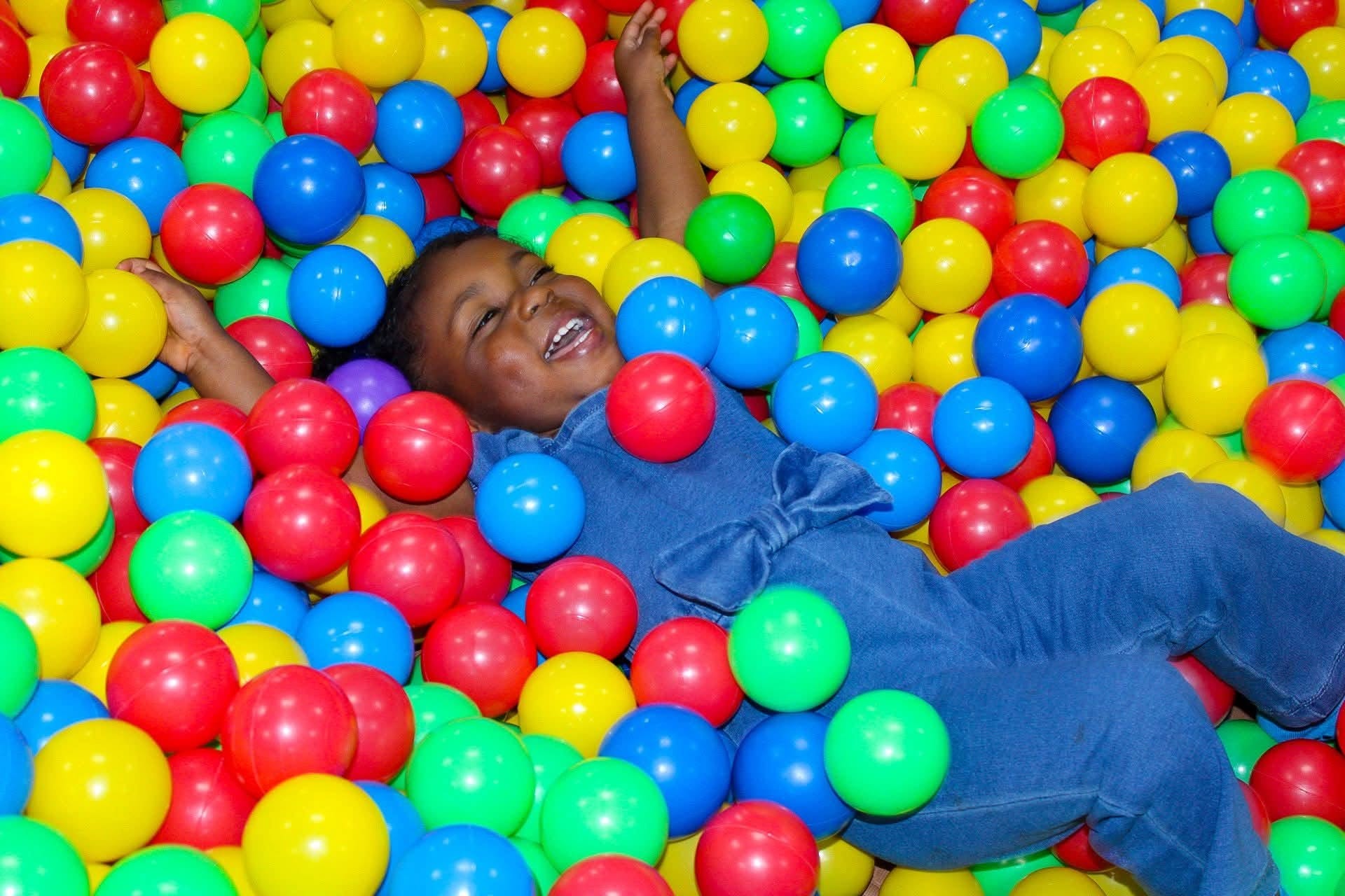Child lying in a colorful ball pit, smiling with arms raised, surrounded by red, yellow, green, and blue balls.
