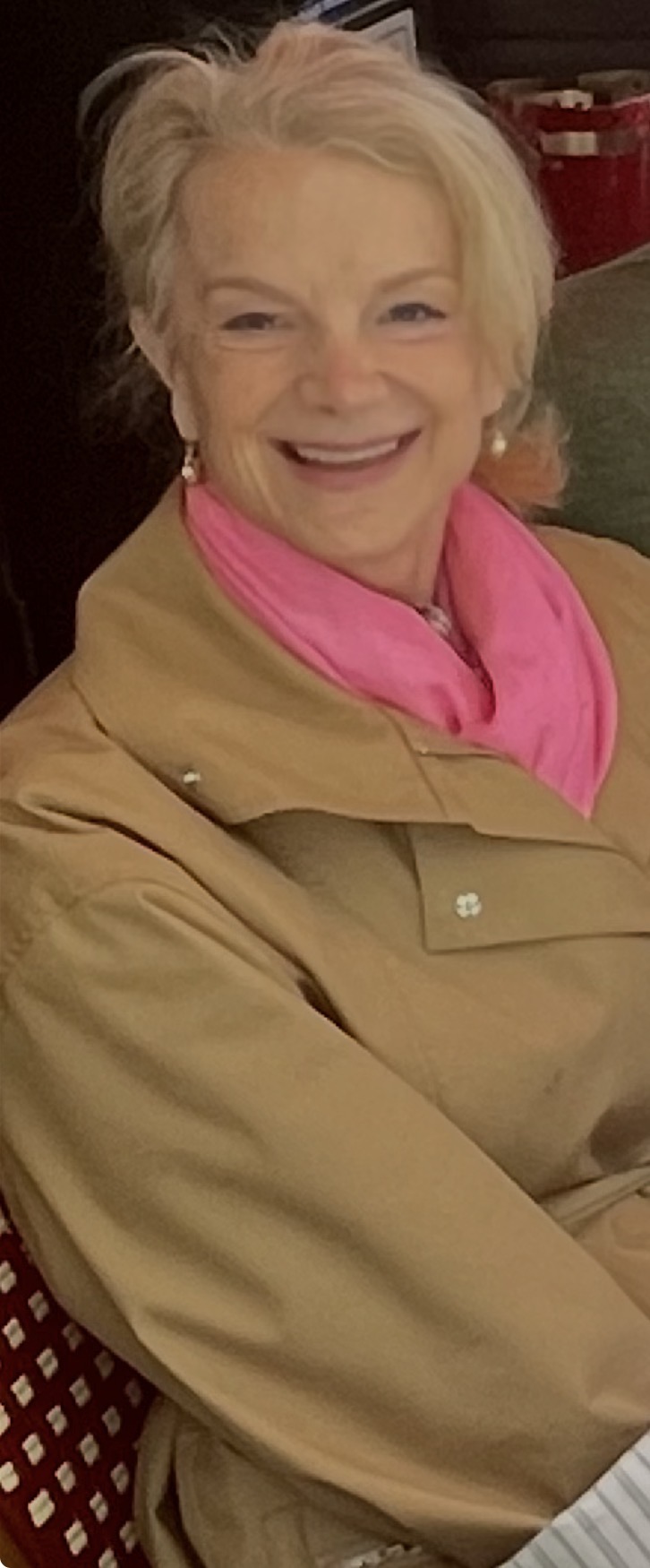 Smiling elderly woman with white hair, wearing a beige jacket and pink scarf, sitting indoors.