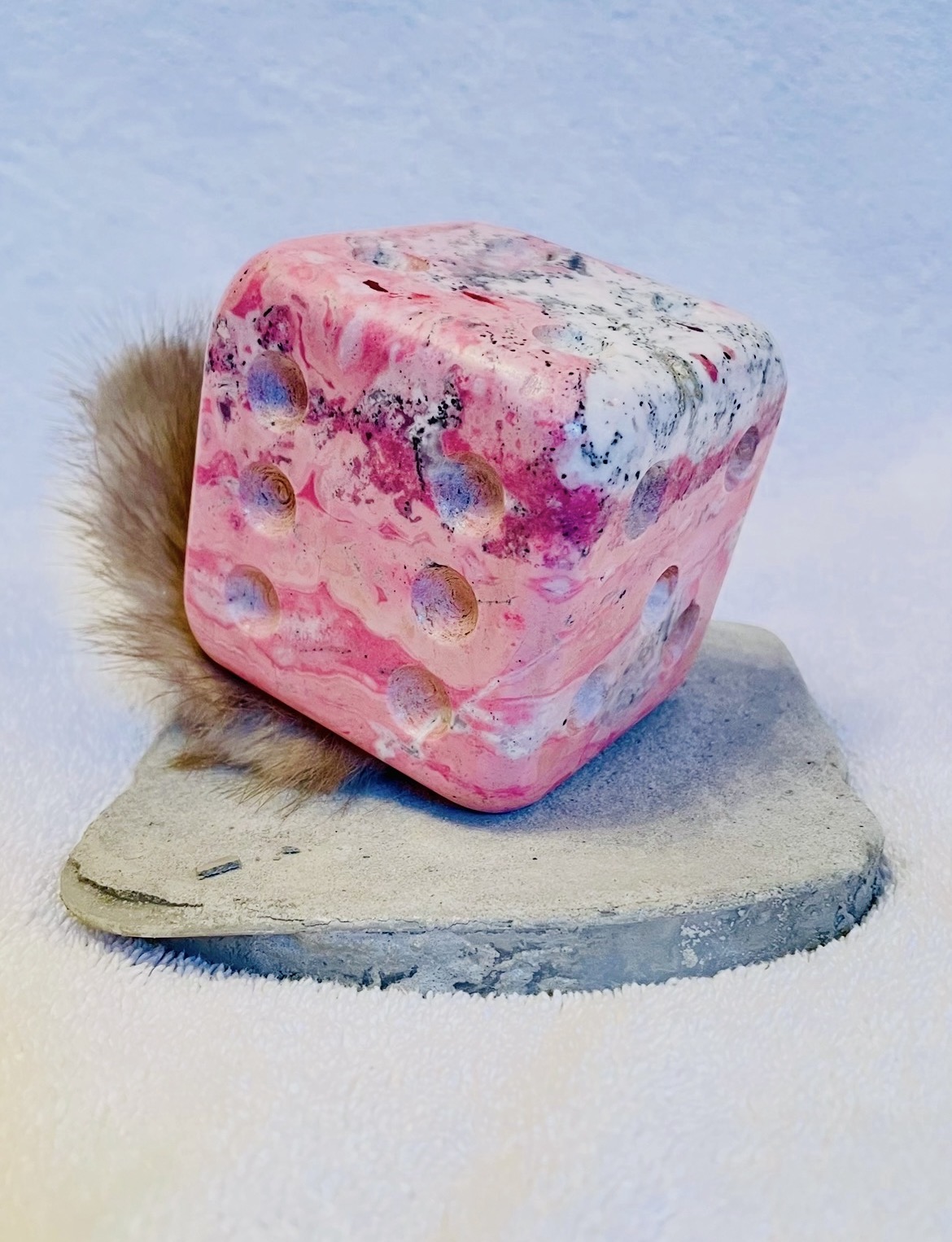 A pink, marbled cube with holes, placed on a gray, irregularly shaped surface, with a furry object behind it.
