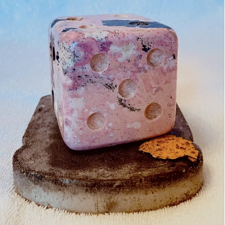A pink and purple marbled die with six circular indentations, resting on a dark, textured surface with a small orange object nearby.