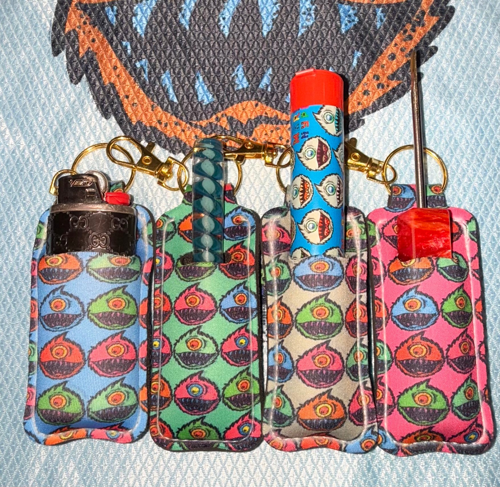 Four colorful lighter cases with eye patterns, attached to a keychain, on a fabric background with a large eye design.