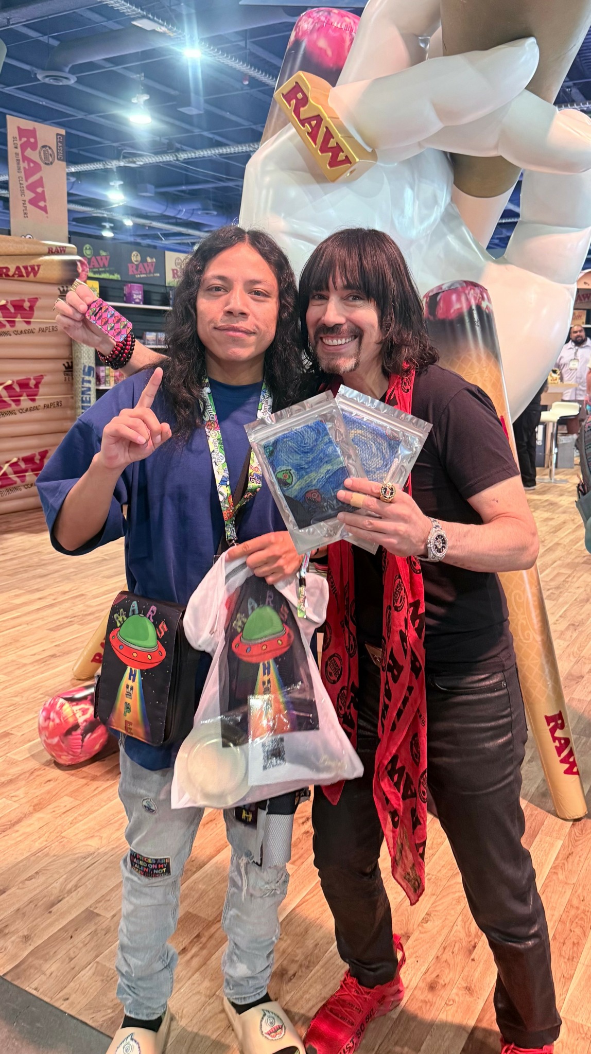 Two women standing together at a convention, holding merchandise and smiling, with a large inflatable figure in the background.
