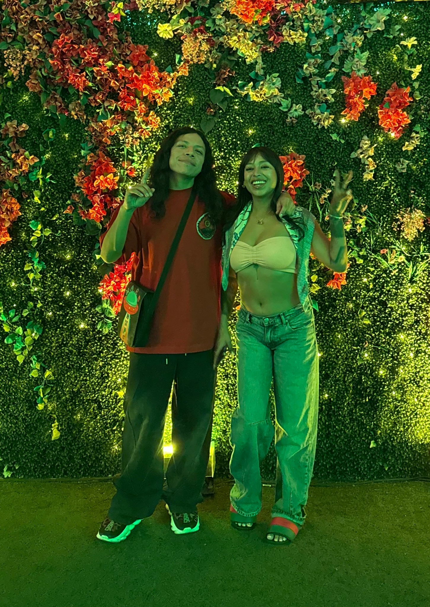 Two women stand in front of a colorful floral wall, smiling and making peace signs, one in a red shirt and the other in a yellow top.