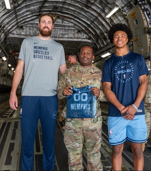 Three people standing inside a spacecraft, one in military uniform holding a Memphis Grizzlies bag, two in casual sportswear, smiling.