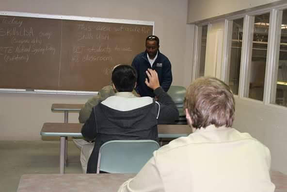 A classroom with a teacher standing in front of a blackboard, students seated facing the teacher, one student raising a hand.