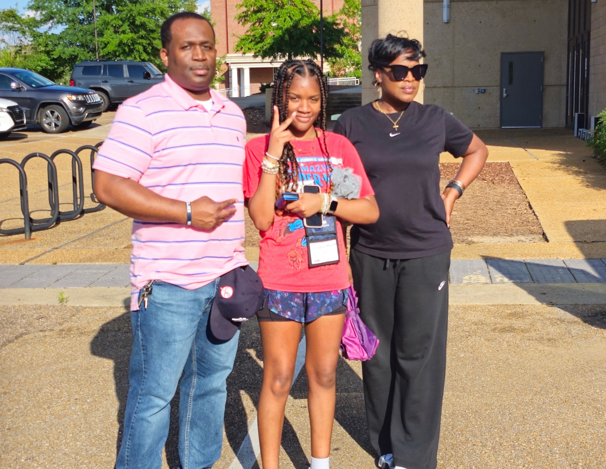 Three people standing outdoors, smiling, with cars and a building in the background.